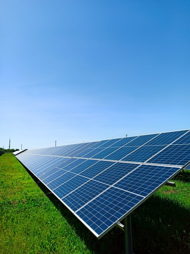 A row of solar panels stand on a green field, against the blue sky. A symbol of clean energy, concern for the environment, a concept.